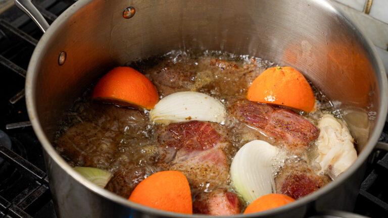 Pork butt, onion, oranges, and spices cooking in a large stainless pot.